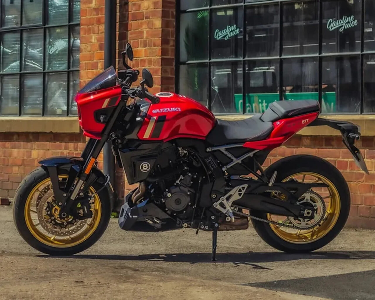 Red Suzuki sport motorcycle with gold wheels parked on a street in front of a brick building with large windowed storefronts, side stand down.