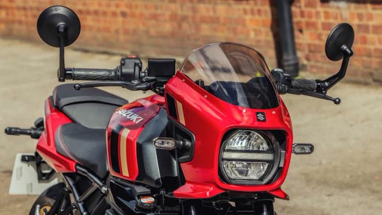 Front view of a red Suzuki motorcycle with a windshield and dual headlights against a brick wall background, shows racing stripes on the side fairing.