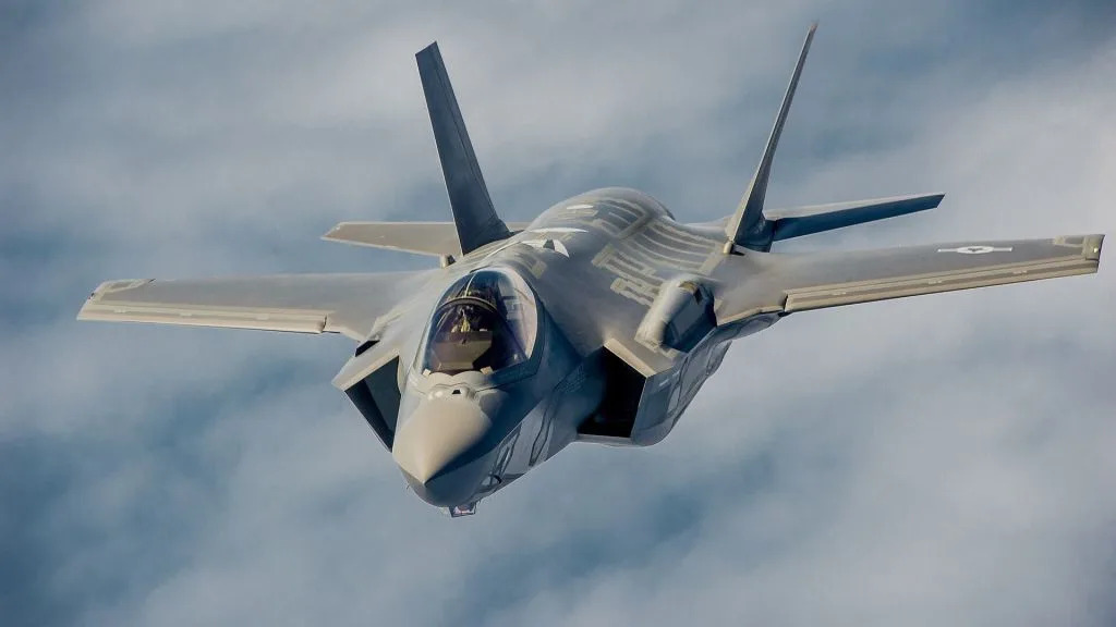 Front-view image of a stealth fighter jet flying toward the camera through a cloudy blue sky, wings spread wide.