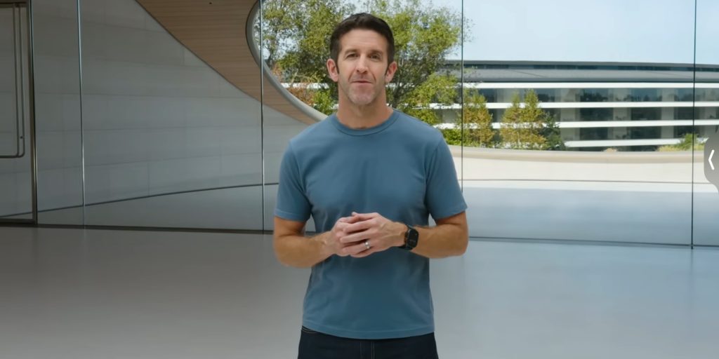 Man in a blue t-shirt stands indoors, hands clasped, wearing a black smartwatch in a modern glass-walled space.