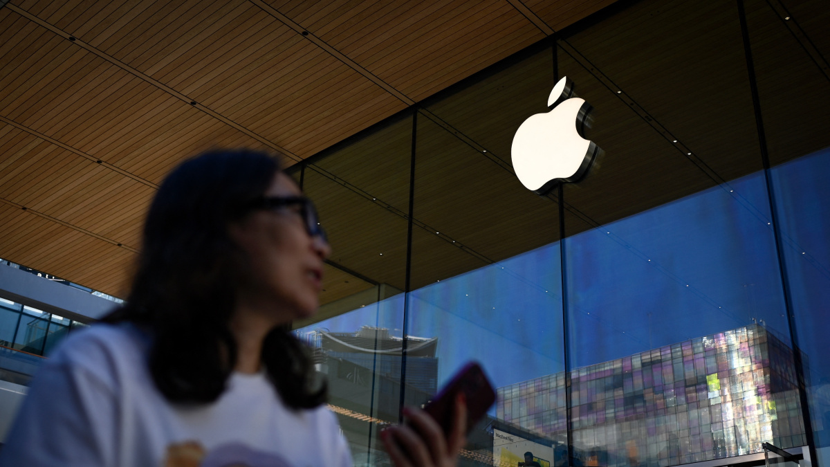 Blurred woman in the foreground outside an Apple Store, with a large white Apple logo on the glass wall behind her.