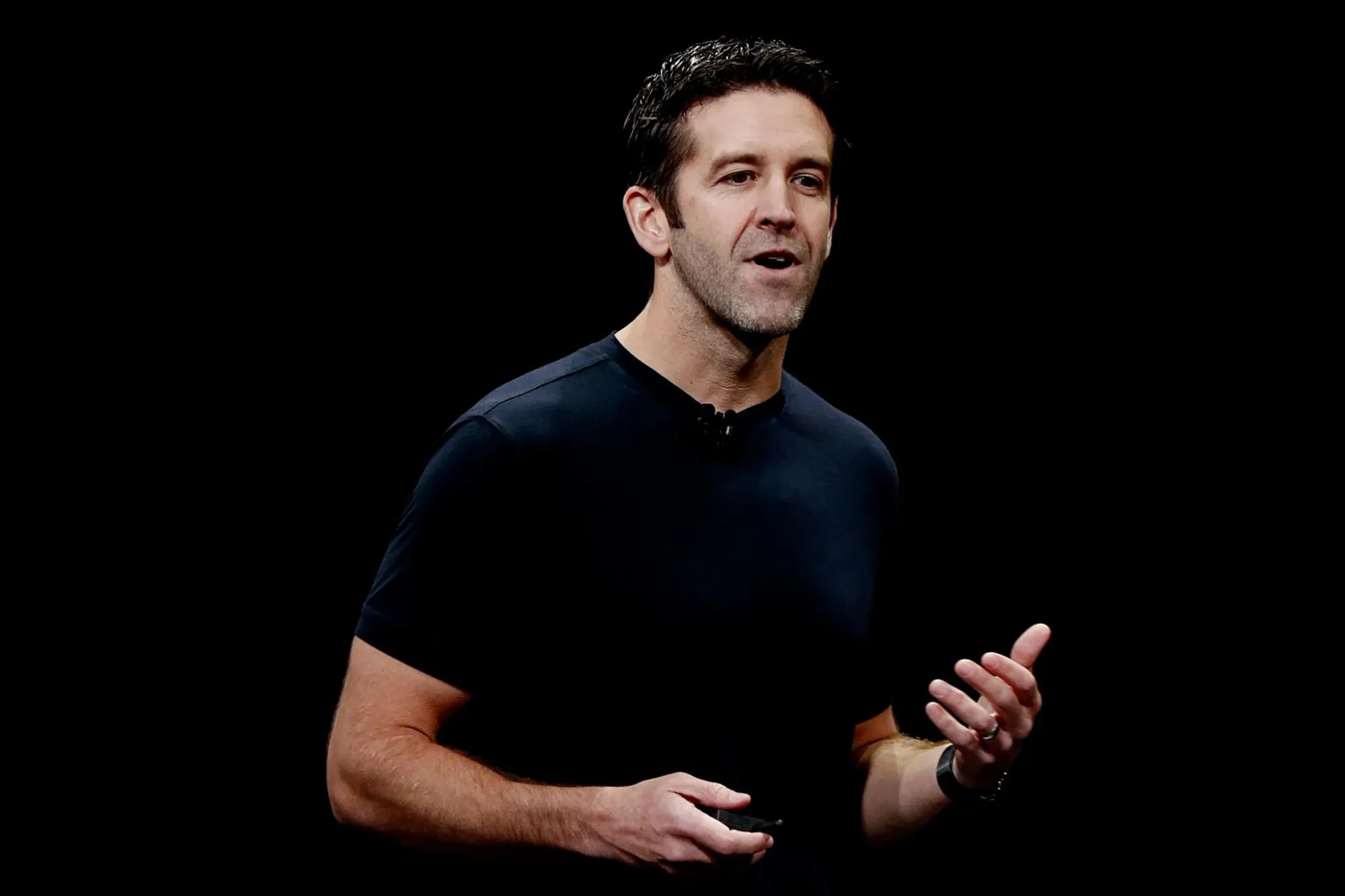 Man in a dark T-shirt speaking on stage, gesturing with his hands against a black background