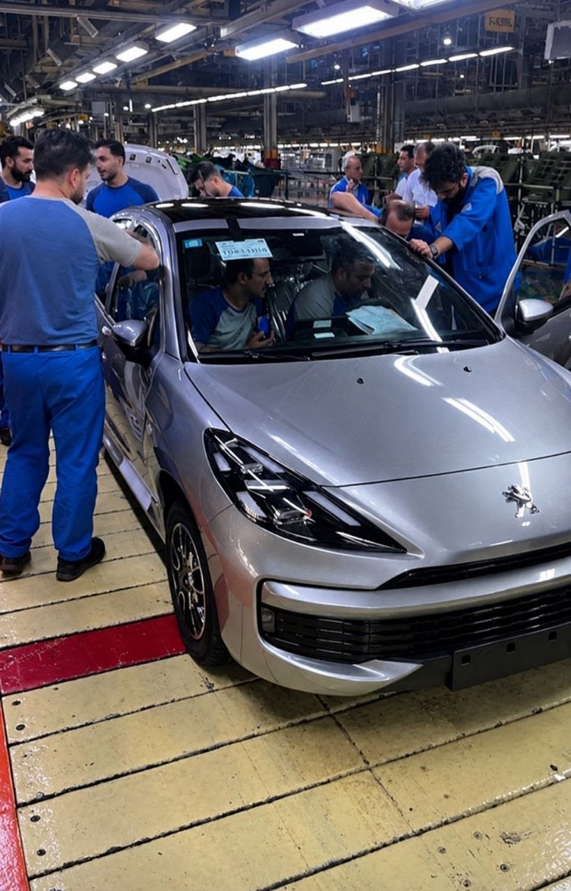 Silver Peugeot hatchback on an automotive assembly line with workers in blue uniforms inspecting the interior through the open doors.