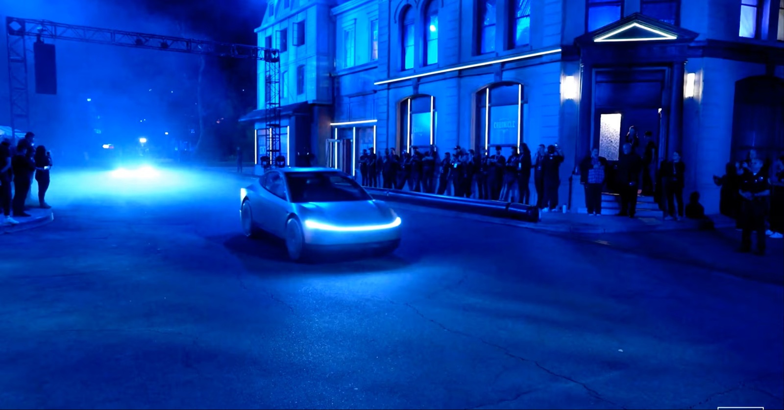 Futuristic blue-lit car drives down a city street at night as a crowd watches from the sidewalk and a stage rig overlaps in the background.
