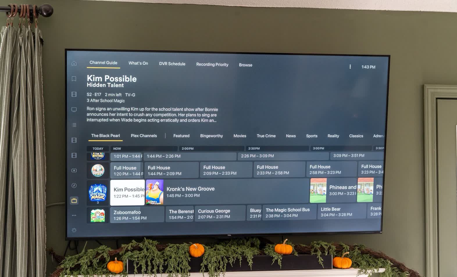 Wall-mounted TV displaying a channel guide with a timeline and program names, plus autumn pumpkin decor on the mantle below