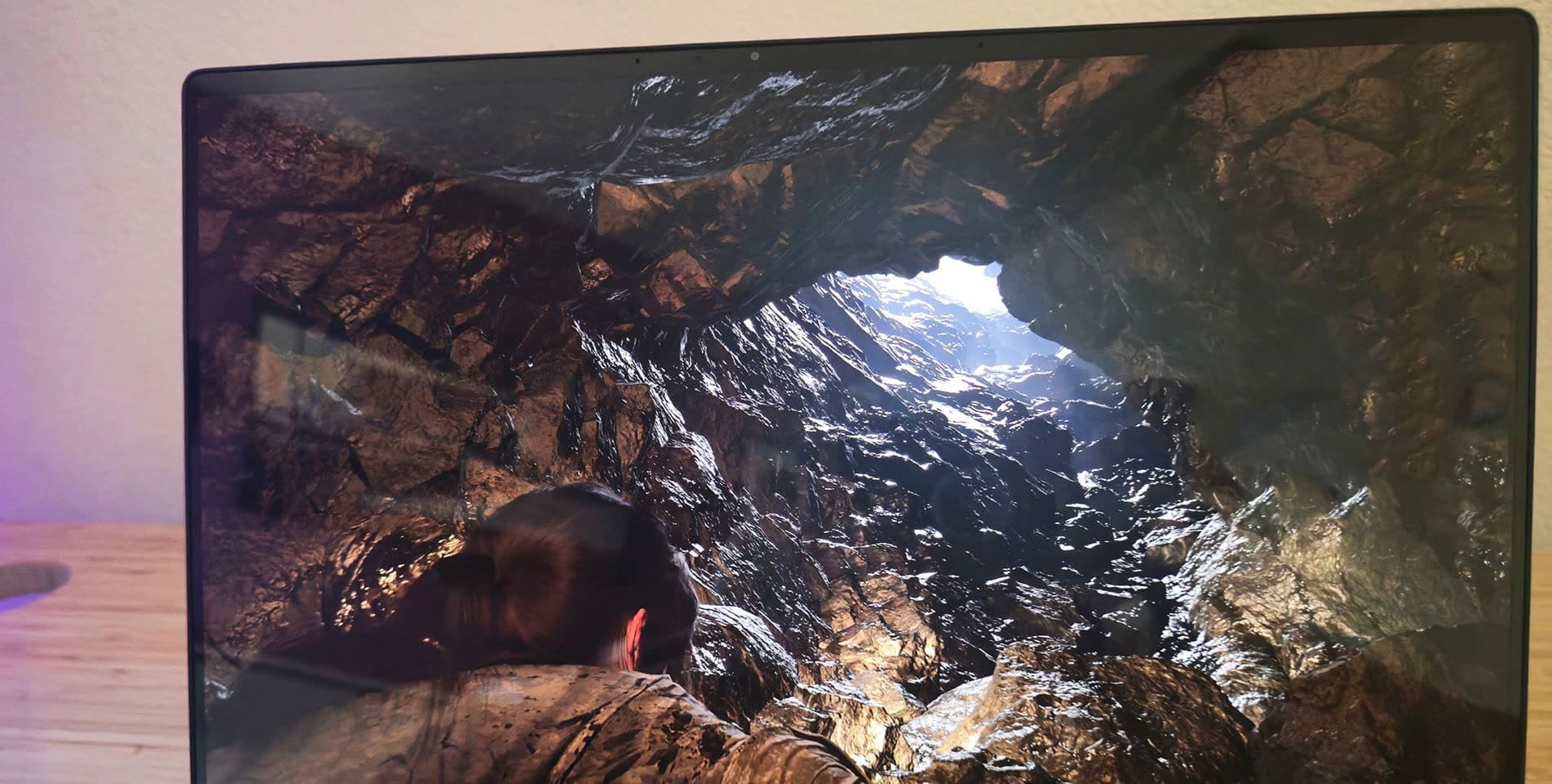 Close-up of a computer monitor showing a rocky cave interior, with a person in a brown jacket at the bottom foreground.