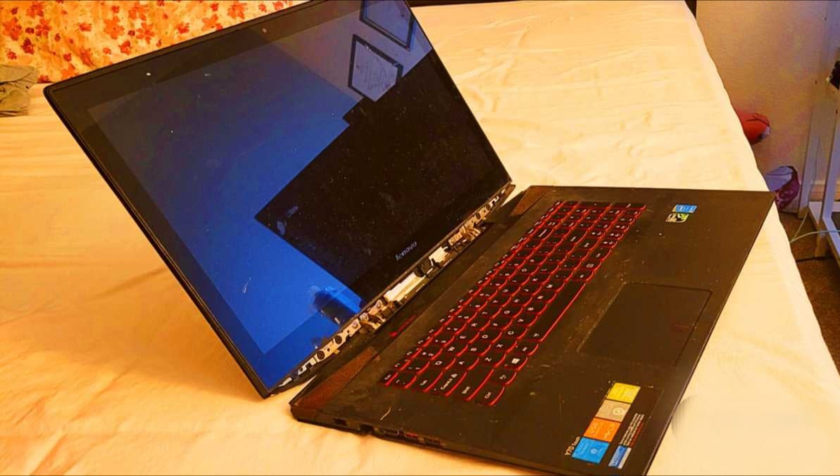 Open Lenovo laptop with red-backlit keyboard resting on a bed, screen angled back and damaged hinge area visible.