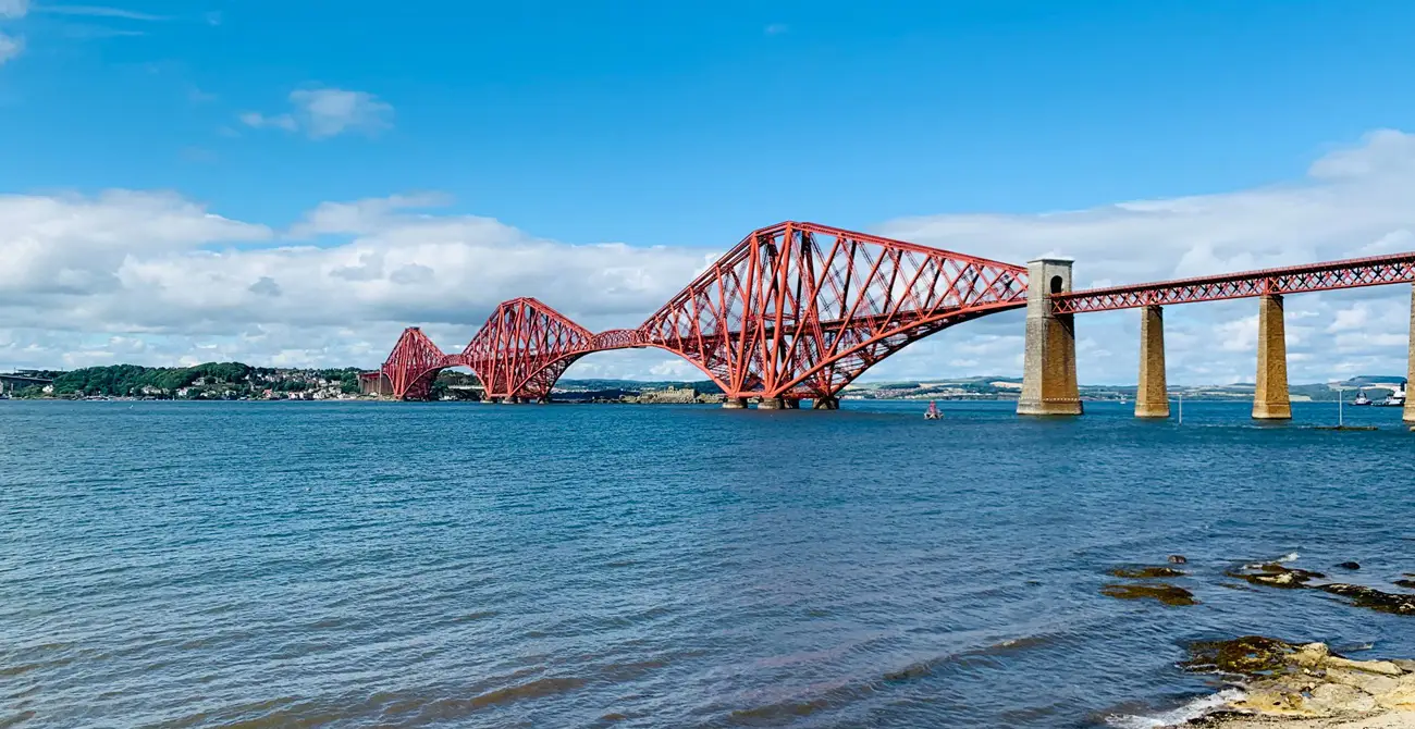 Red cantilever railway bridge crossing blue water, with a distant shoreline and blue sky.