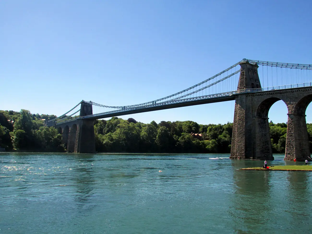 Suspension bridge spanning a wide river with stone towers, flanked by trees on a sunny day.