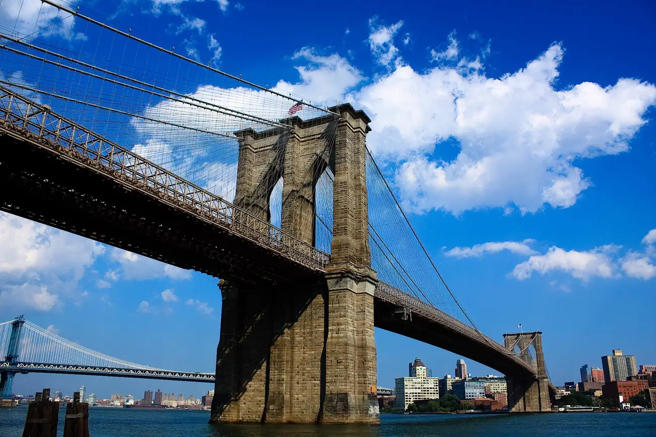 Brooklyn Bridge with stone towers, suspension cables, and a blue sky over the East River, with Manhattan skyline in the background