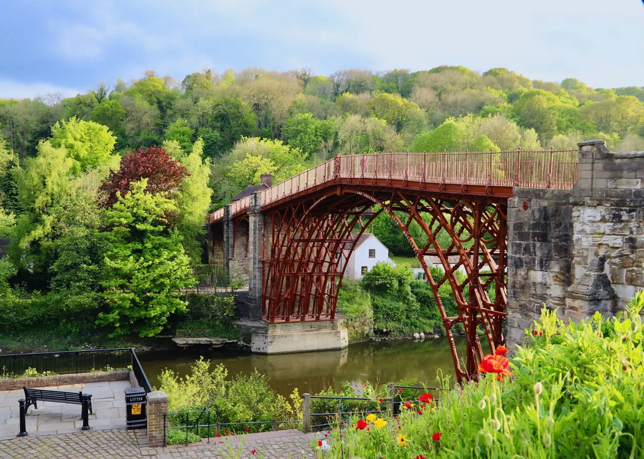Red wrought-iron lattice bridge crossing a calm river, with lush green trees and a small white house along the bank. foreground colorful flowers.