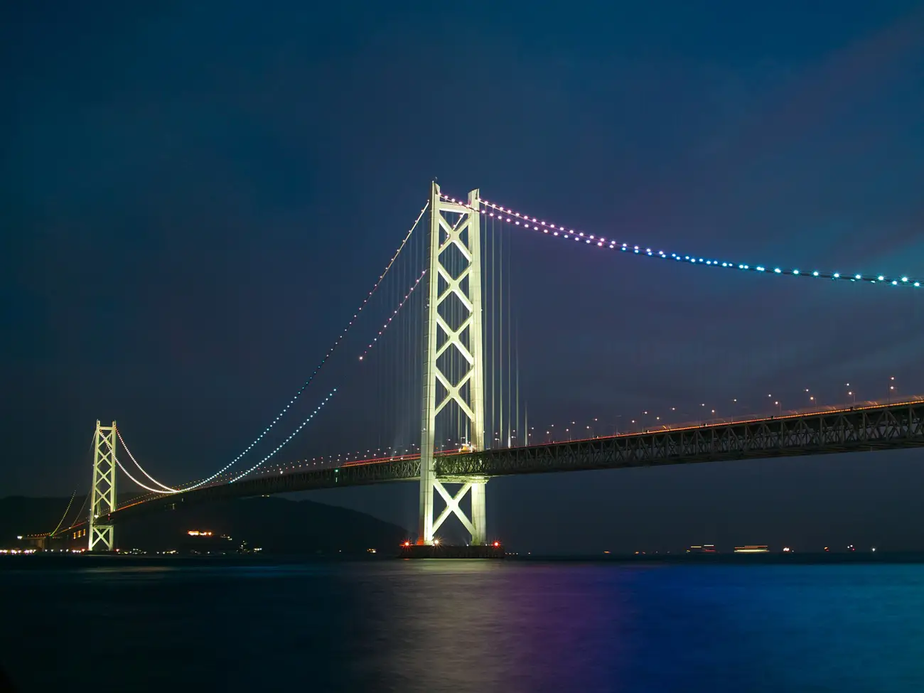 Night view of a suspension bridge lit with colorful LEDs, spanning calm water with reflections below.