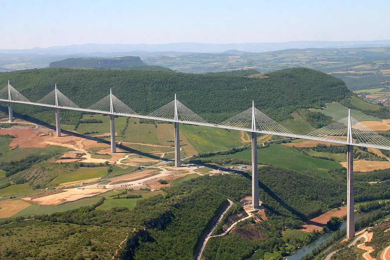 Cable-stayed bridge with tall white pylons spanning a green valley over a winding river. Reflective cables fan from each pylon to the deck amid rural fields and hills.