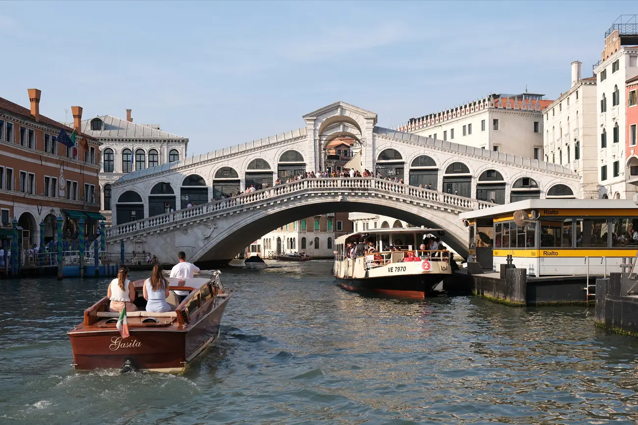 Wooden tour boat glides on a Venetian canal under the arches of the Rialto Bridge, with people aboard and city buildings beside.