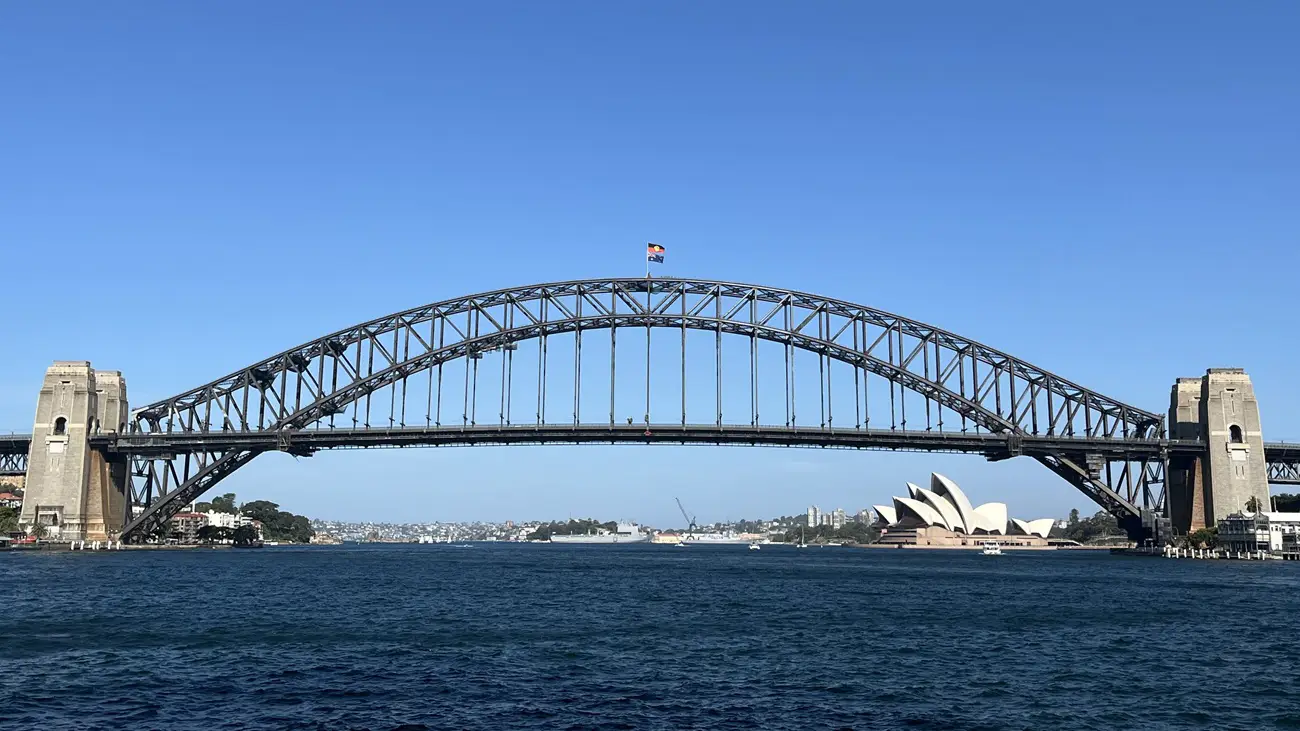 Sydney Harbour Bridge arches over blue water with the Opera House visible on the right under a clear sky.