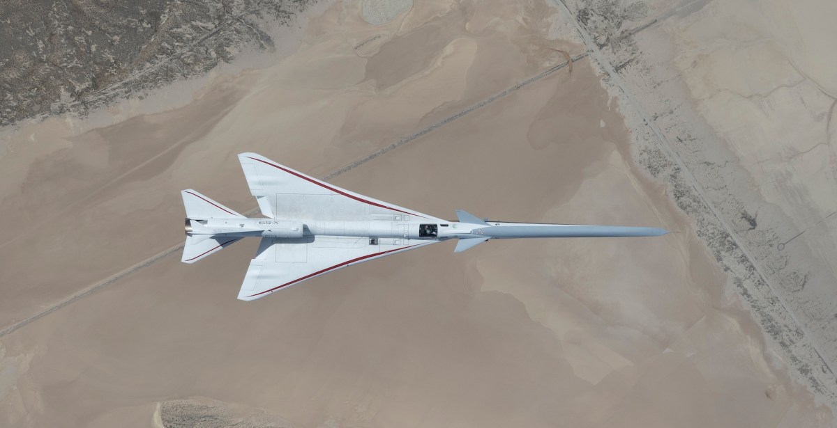 NASA’s white, blue, and red X-59 research aircraft flies over the Mojave Desert near NASA’s Armstrong Flight Research Center in Edwards, California, during its third flight. The aircraft is centered in the frame, viewed from above with the canopy, both wings, NASA and X-59 logos visible, and the landing gear down with one wheel visible. Below, rugged terrain transitions into the smooth texture of a dry lakebed as the aircraft, nearly 100 feet long, flies toward the right side of the frame.