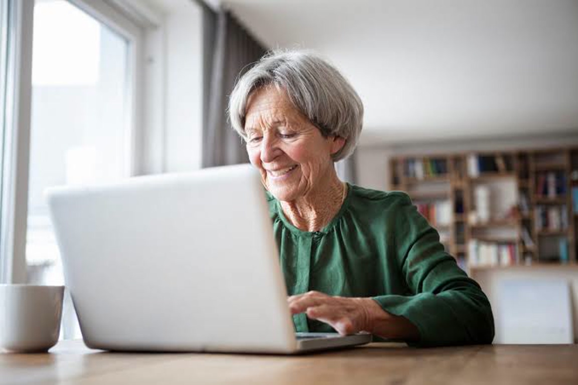 Smiling elderly woman using a laptop at a wooden table by a window