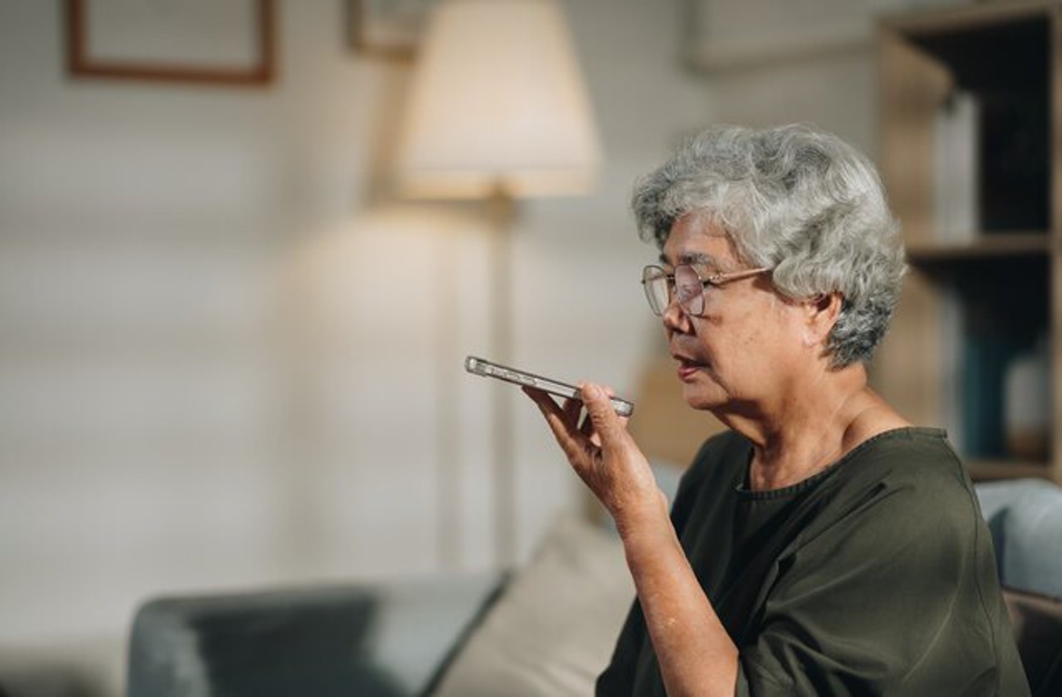 Older woman with gray hair and glasses sits on a sofa at home, holding a digital thermometer to her mouth.