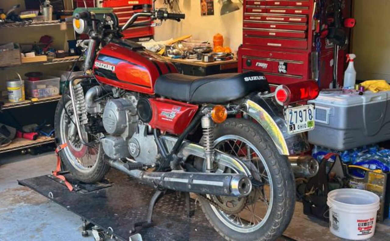 Red Suzuki motorcycle on a maintenance lift in a cluttered home garage with tool chests and shelves nearby