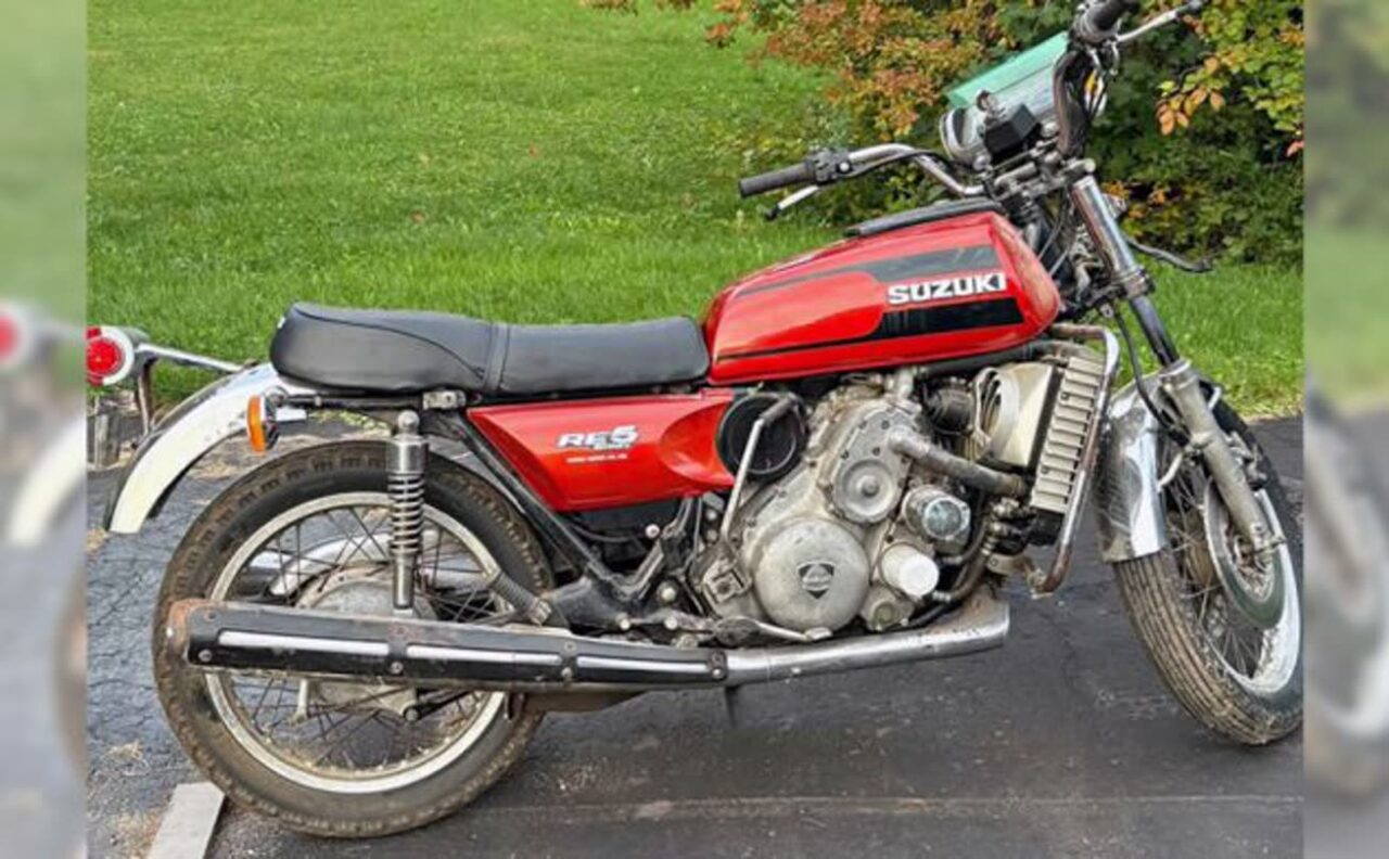 Red vintage Suzuki motorcycle parked on a driveway with a black seat and chrome exhaust, green grass in the background.