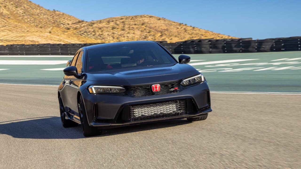 Front three-quarter view of a dark gray Honda Civic Type R driving on a race track, with desert hills and tire barriers in the background and a blue sky.