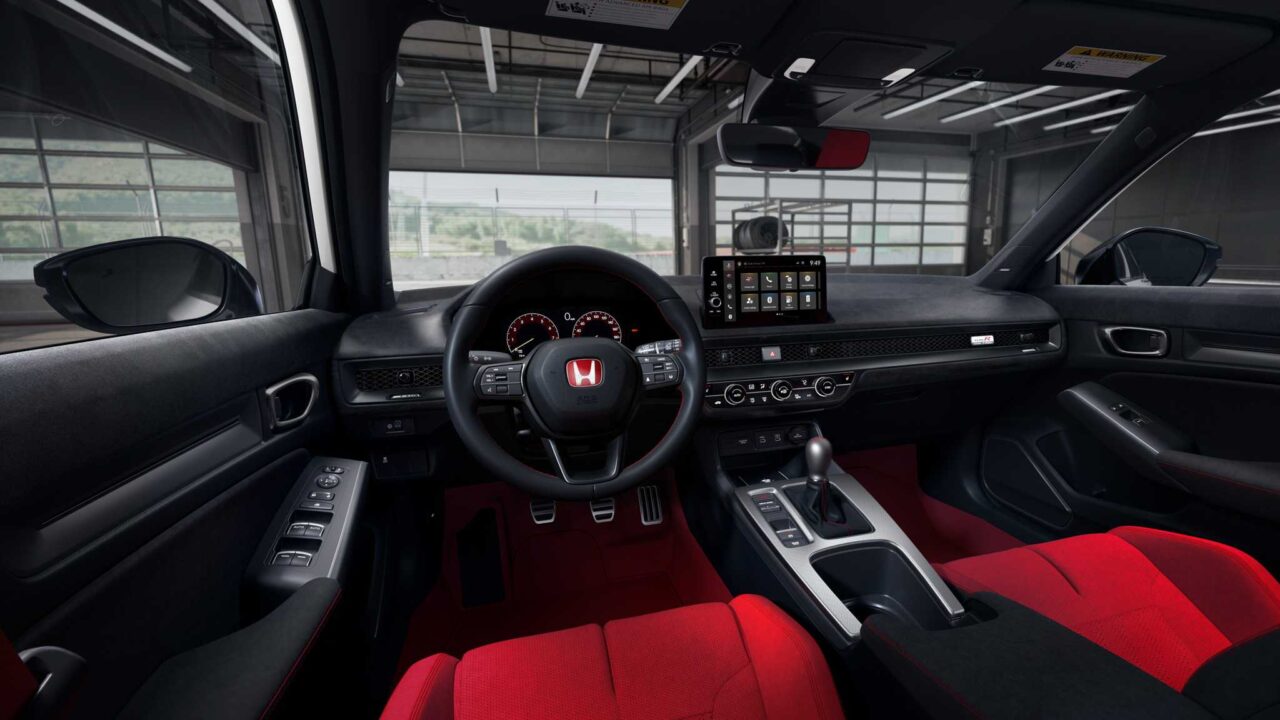 Interior view of a car cockpit with a red upholstered driver and passenger seats, black dashboard, steering wheel, center touchscreen, and gear lever in view.