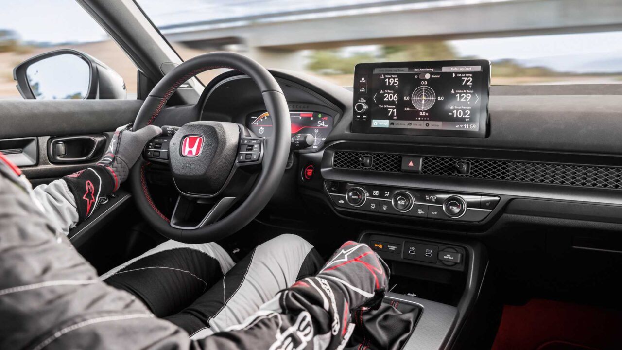 Racing driver seated in a Honda car cockpit, hands on the steering wheel with a digital instrument display ahead.