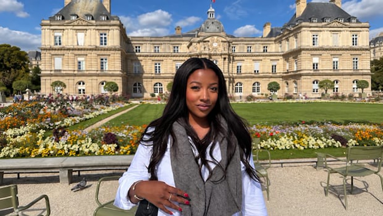 Young woman with long dark hair posing outdoors in front of a grand stone club-style building and a colorful flower garden under a blue sky.