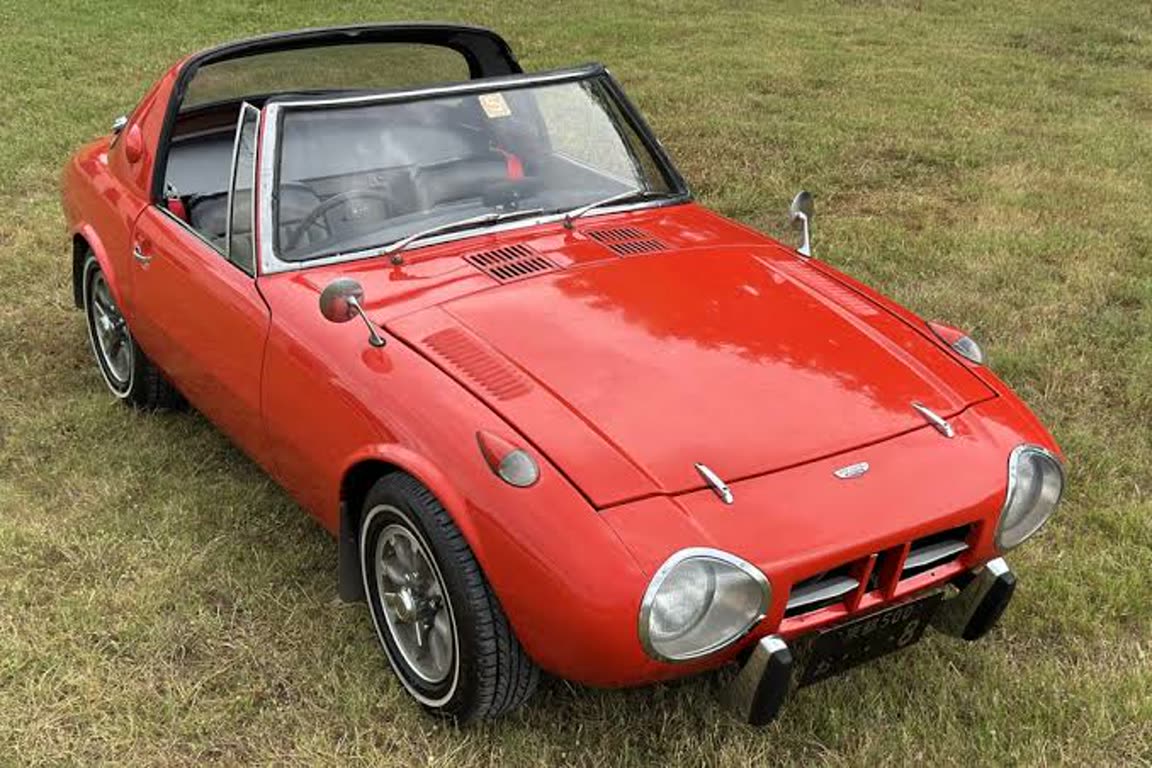 Red vintage convertible sports car parked on a grassy field, two-seat interior visible with soft-top down