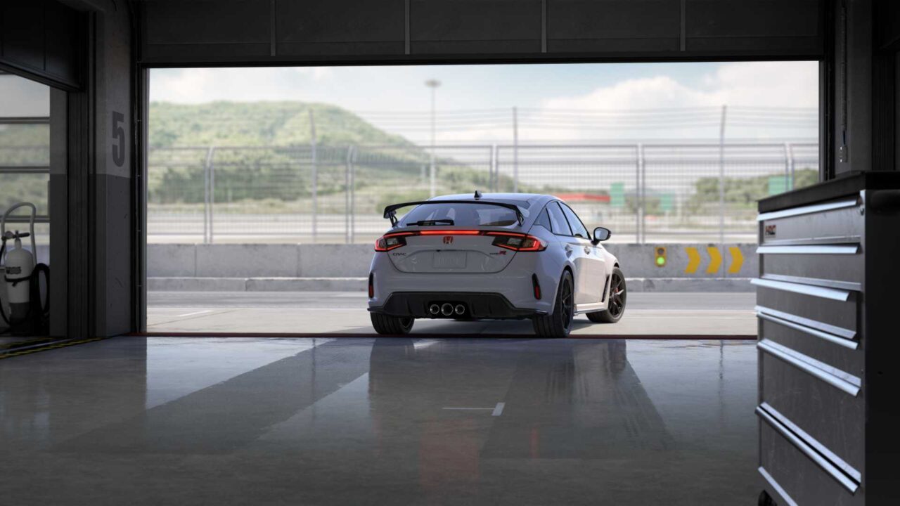 White Honda Civic Type R parked in a dim pit garage, rear view, race track visible outside frame.