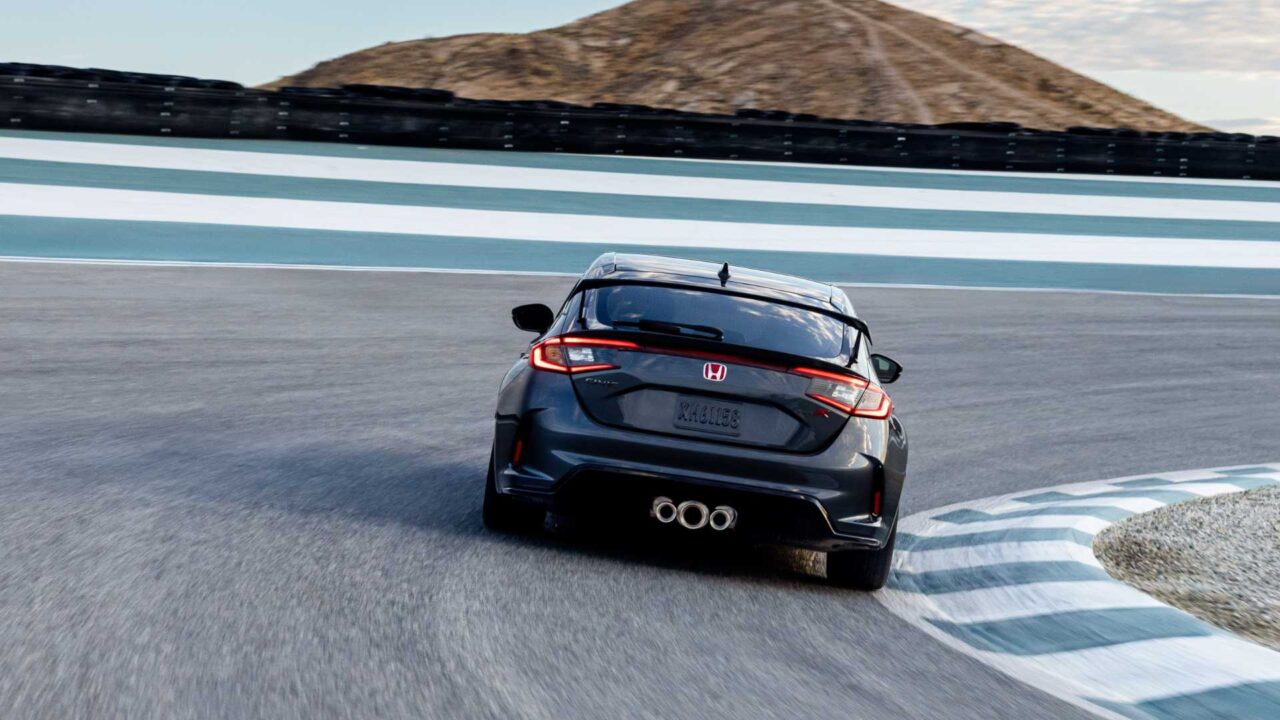 Rear view of a dark Honda Civic hatchback taking a sharp turn on a race track, with blue-and-white barriers and a hill in the background.