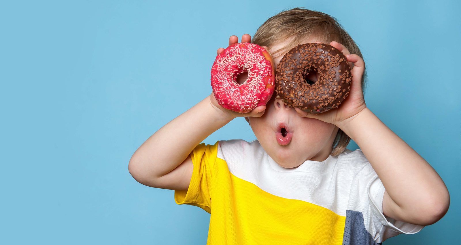 Child playfully holds two frosted donuts in front of eyes against a blue background, making a goofy gaze.