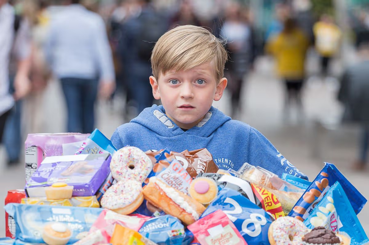 Young boy in a blue hoodie sits behind a large pile of snacks and candy on a busy street, pedestrians blurred in the background.
