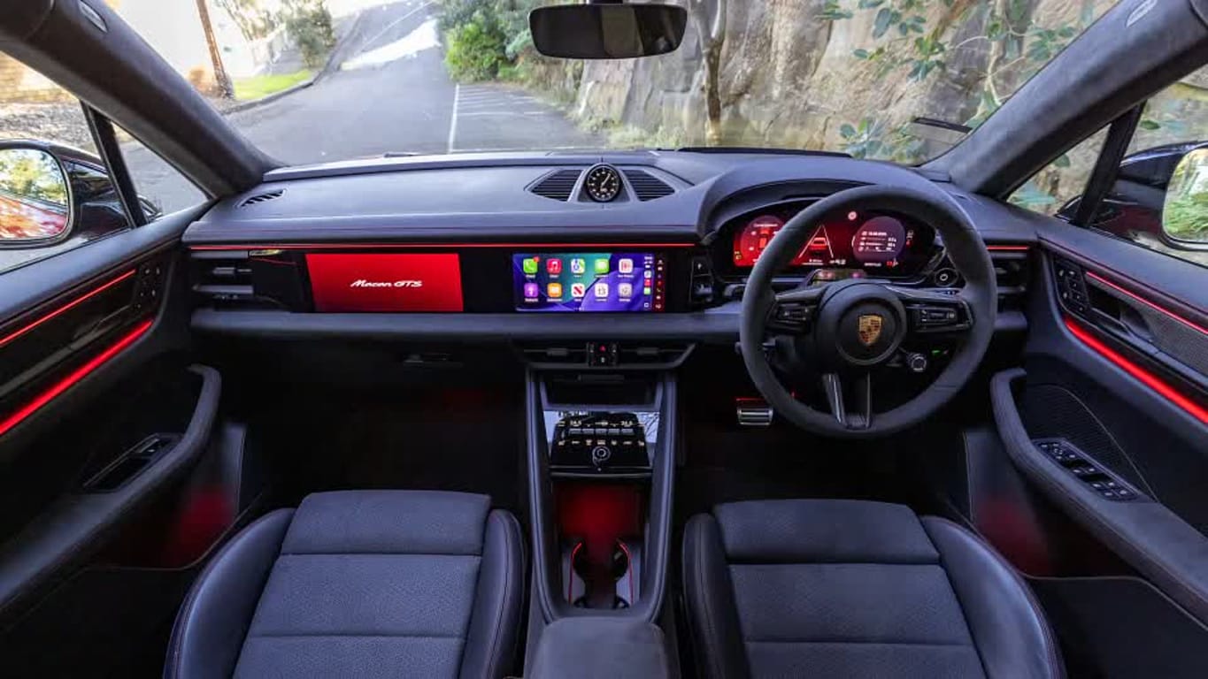 Inside view of a dark Porsche Macan GTS cockpit with red ambient lighting and a wide touchscreen display.