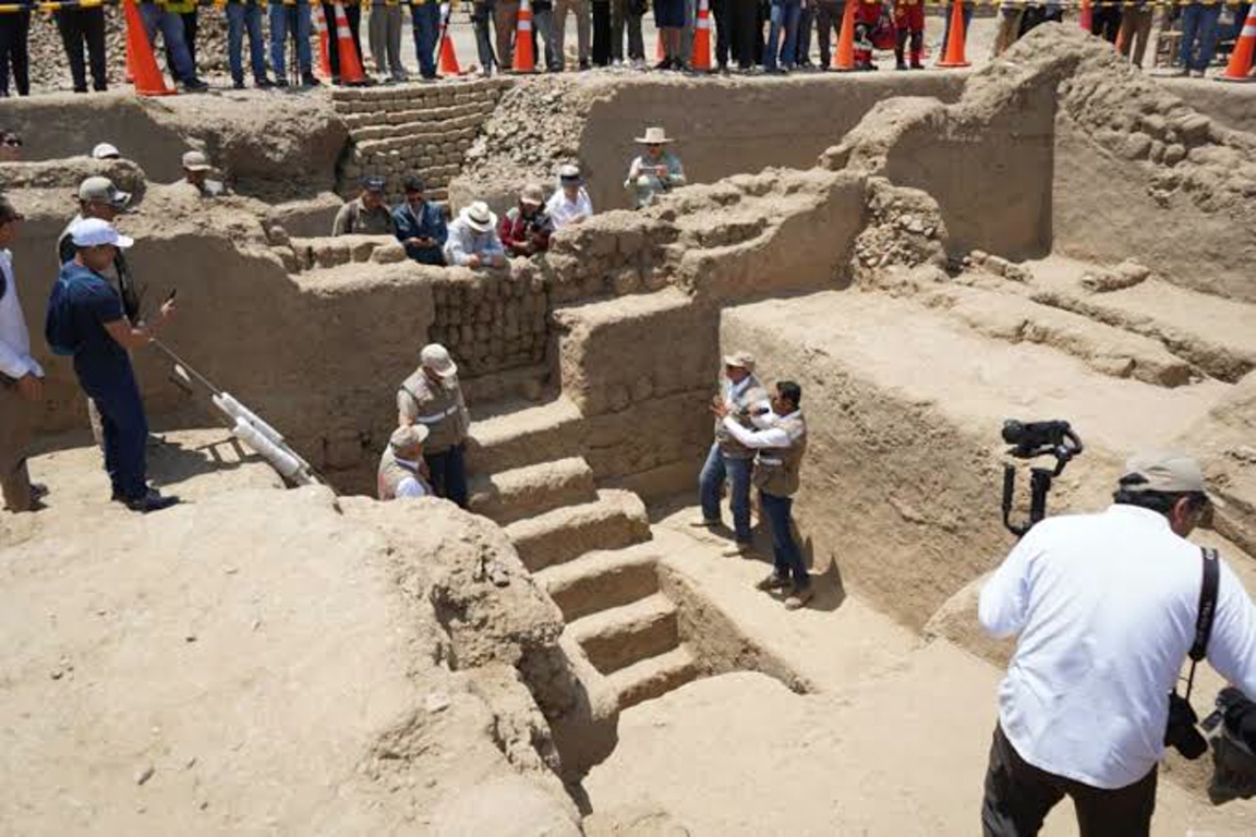 Archaeologists examine exposed adobe walls and a stairway in a sunlit excavation trench, with photographers nearby.