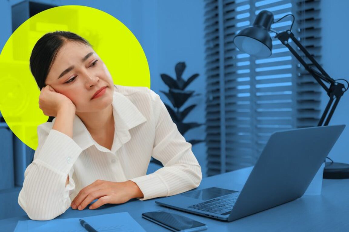 Woman resting her head on her hand at a desk with a laptop, looking fatigued or bored in an office setting.