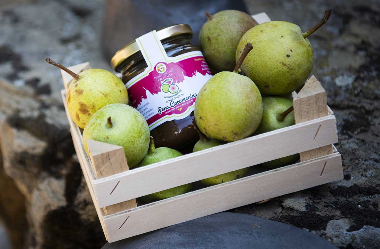 Wooden crate filled with green pears surrounding a jar of fruit jam on a stone surface outdoors.