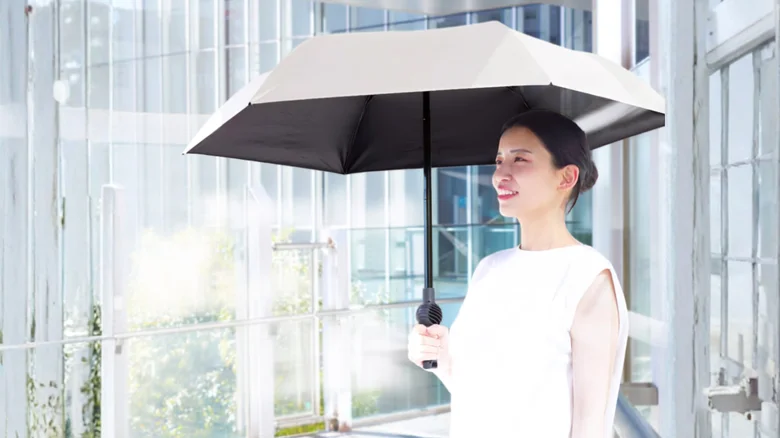 Woman in a white sleeveless top holding a large beige umbrella in a glass-walled atrium, smiling at the camera.
