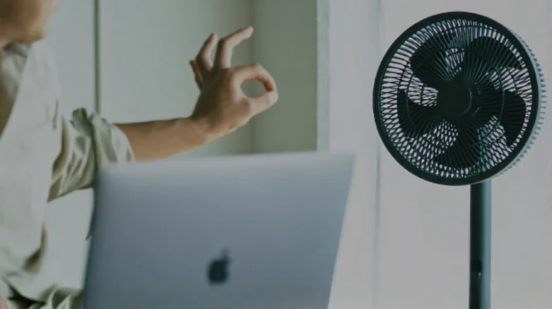 Person working on a laptop while a standing fan sits nearby; hand making an OK gesture in the air.