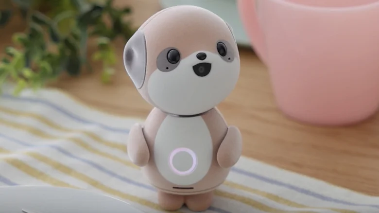 Small beige robotic dog toy with a glowing circular ring on its belly, standing on a striped cloth by a pink mug on a table.
