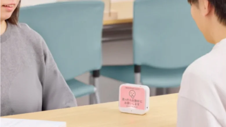 Two people seated and talking at a table with a small pink device on the desk.