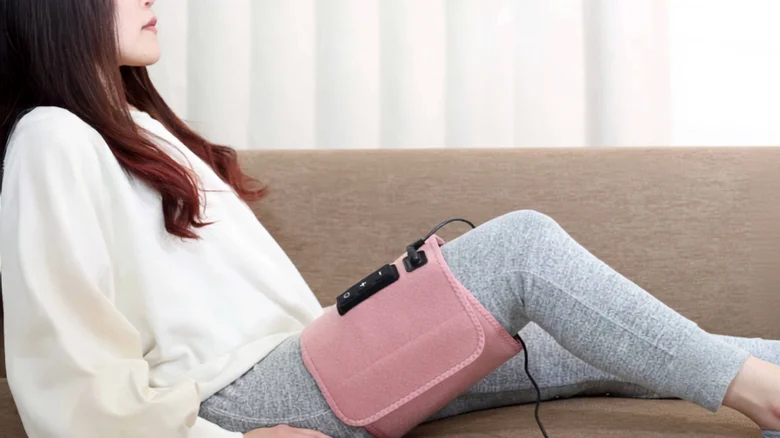 Woman reclining on a sofa wearing a pink leg wrap with a control device and wires, likely a therapeutic muscle stimulation setup on her thigh/leg.