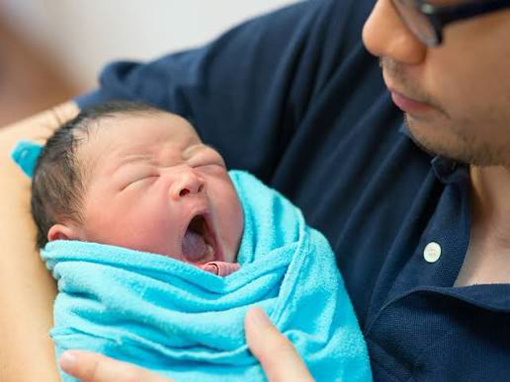 Newborn baby yawning while swaddled in a bright blue blanket, being held by an adult man wearing a dark shirt and glasses.