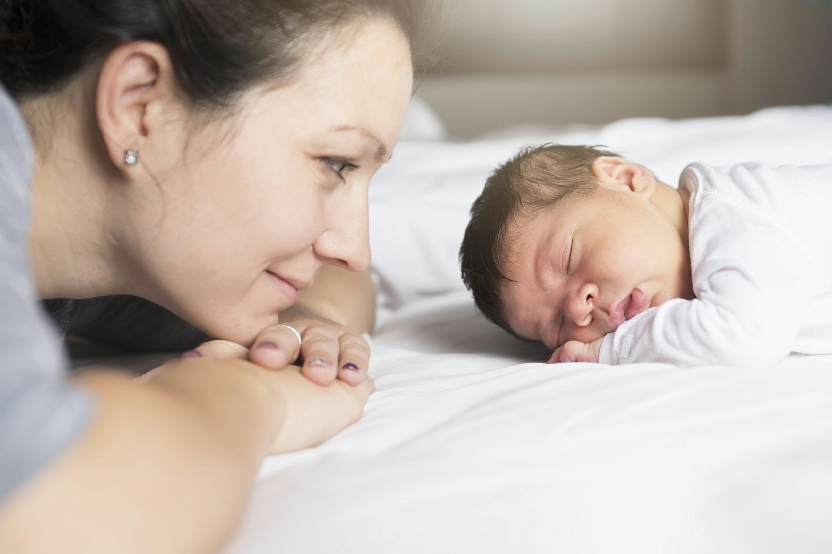 Mother leaning over and smiling at a sleeping newborn on a white bed (tender moment).