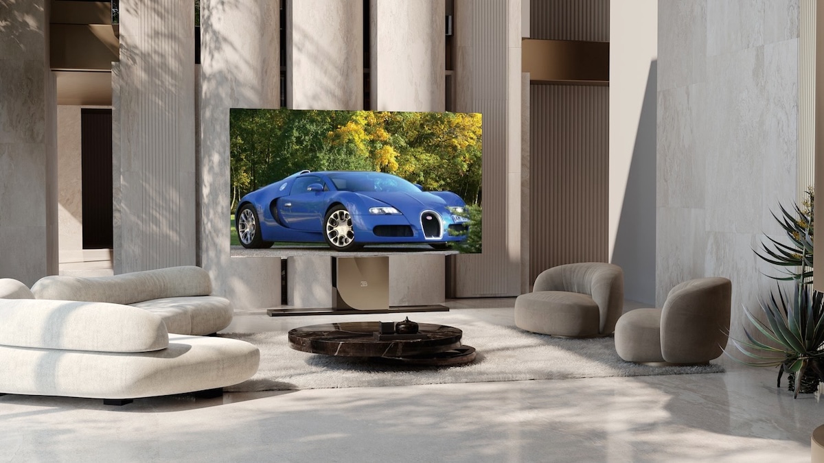 Modern living room with a large flat-screen TV showing a blue sports car, surrounded by beige sofas and a round coffee table.