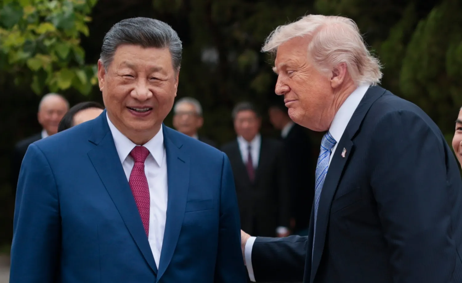 Xi Jinping and Donald Trump in conversation at an outdoor formal event, Xi smiling in a blue suit and red tie while Trump leans in wearing a dark suit and blue tie.