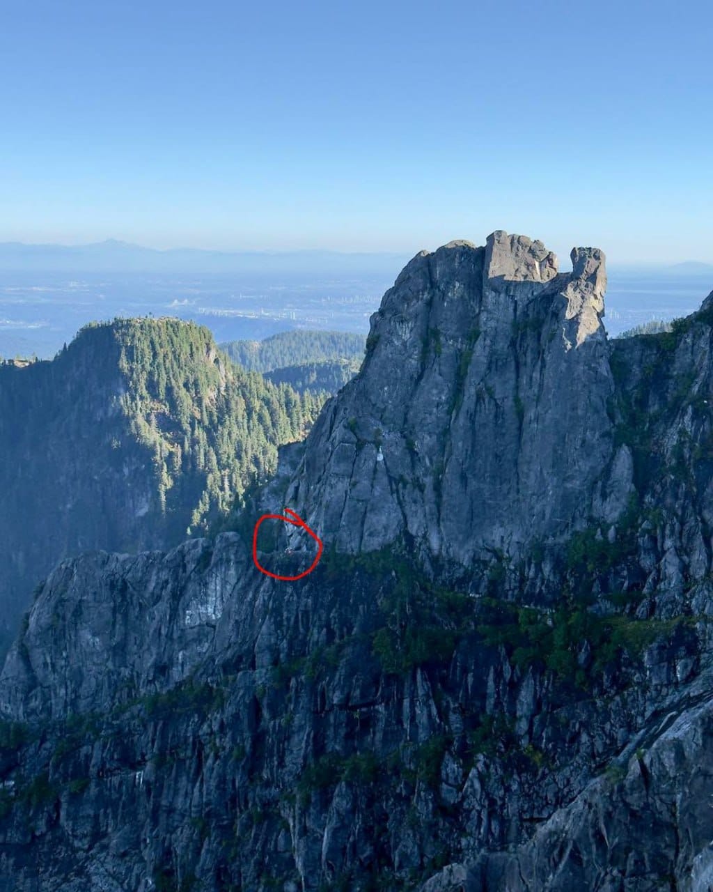 Jagged mountain ridge with steep gray rock faces, forested slopes below, and a distant blue horizon under a clear sky; a small silhouette on a narrow ledge circled in red.