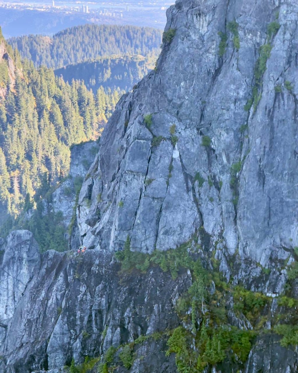 Two hikers on a narrow rock ledge beside a steep granite cliff overlooking a forested valley and distant city on the horizon