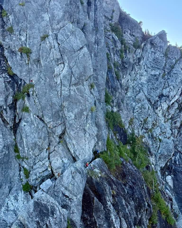 A steep gray rock cliff with small evergreen trees; climbers in red and dark clothing navigate a narrow ledge from an aerial perspective.