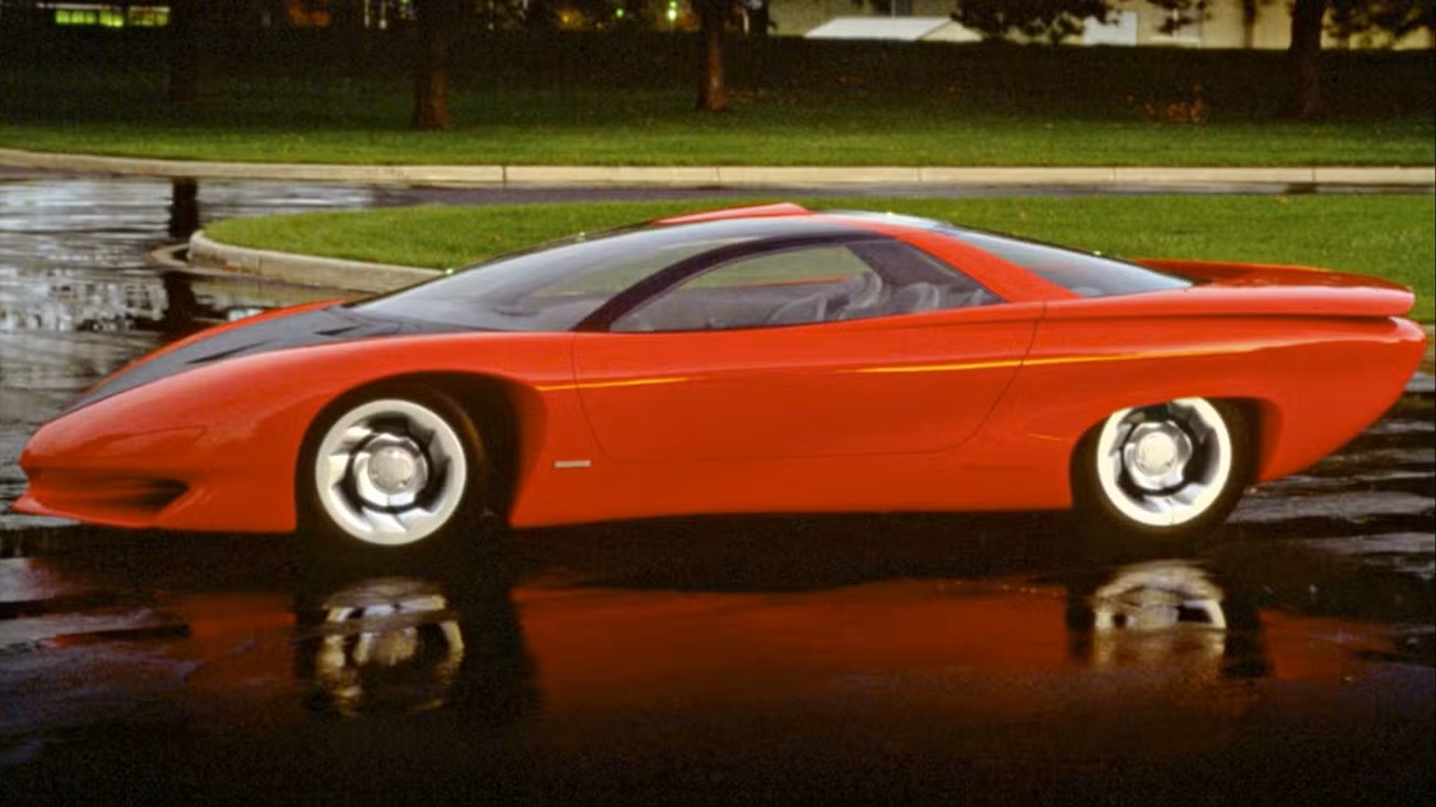 Red futuristic sports car parked on a wet pavement with reflection, green grass and trees in the background.